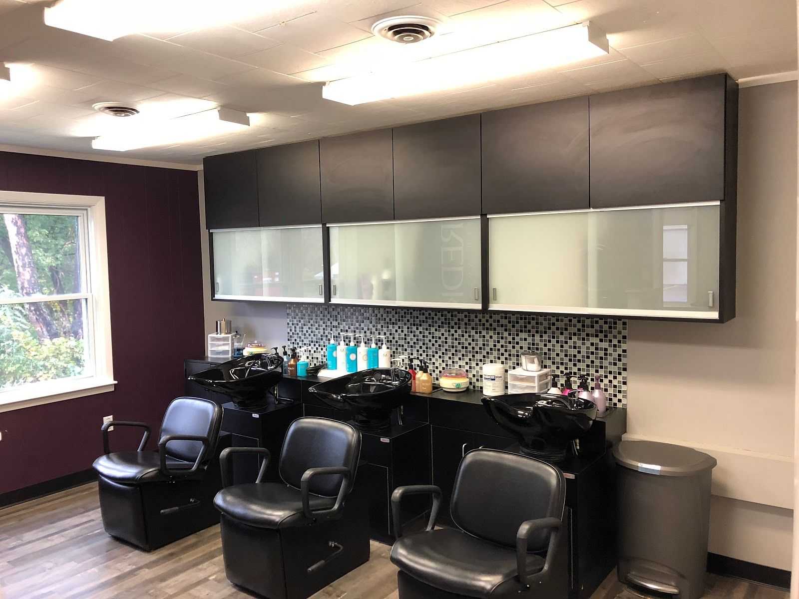 Modern hair salon with three styling chairs, wash basins, and haircare products on shelves.