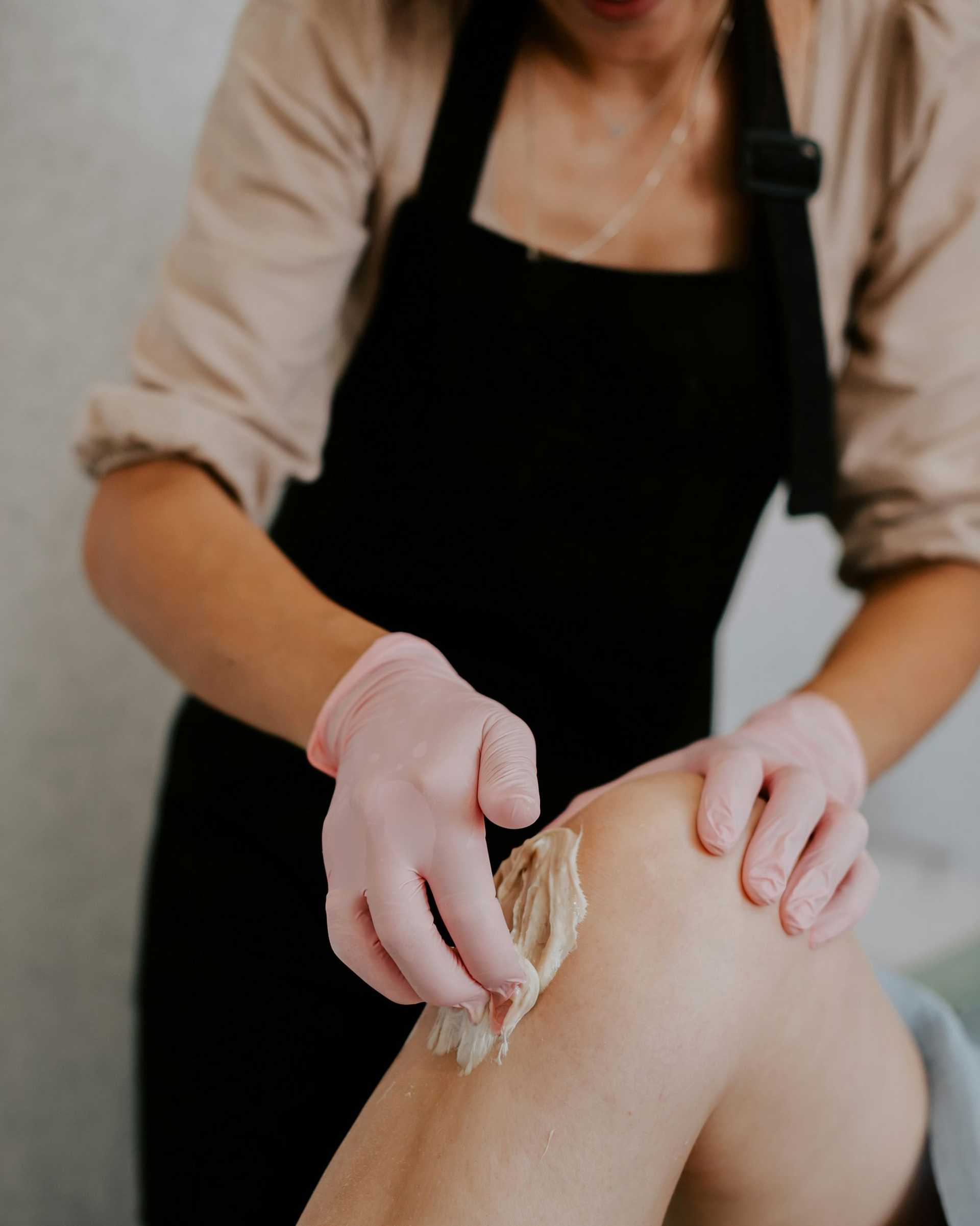 Aesthetician performing waxing on a client's leg wearing gloves.